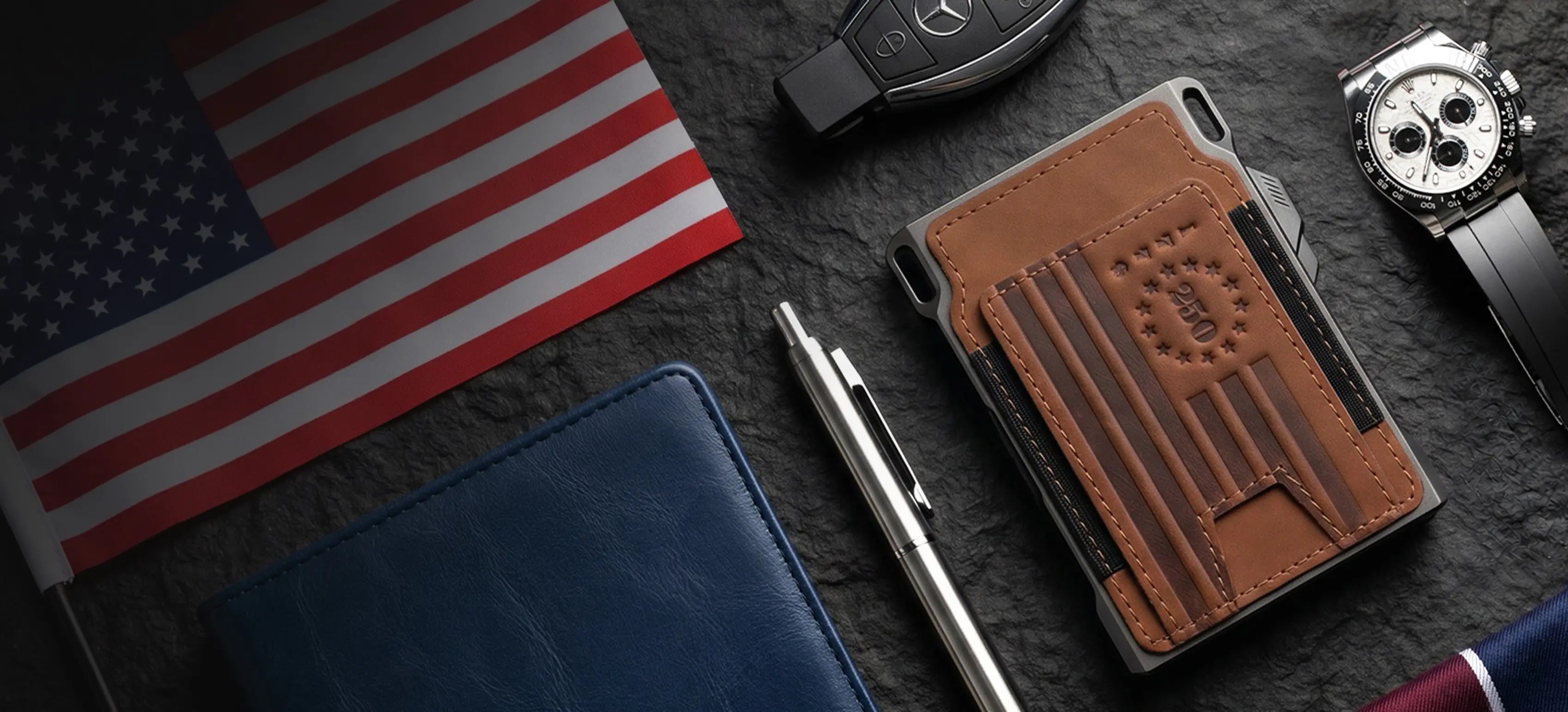 A brown leather card holder wallet with an embossed American flag design and a debossed 'Patriot' logo, styled in a flat-lay arrangement on a dark textured surface alongside an American flag, a blue wallet, a silver pen, a Mercedes key fob, and a wristwatch.