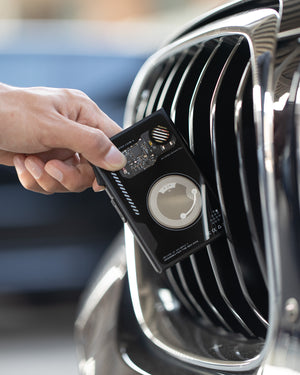 Black wallet held near a car grille, with the back side facing outward and internal circular coil visible.